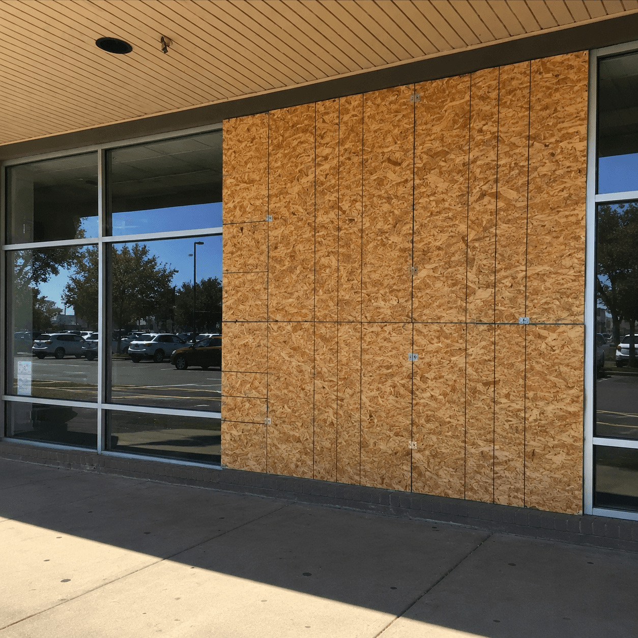 Storefront windows partially covered with plywood boards, reflecting a parking lot and blue sky.