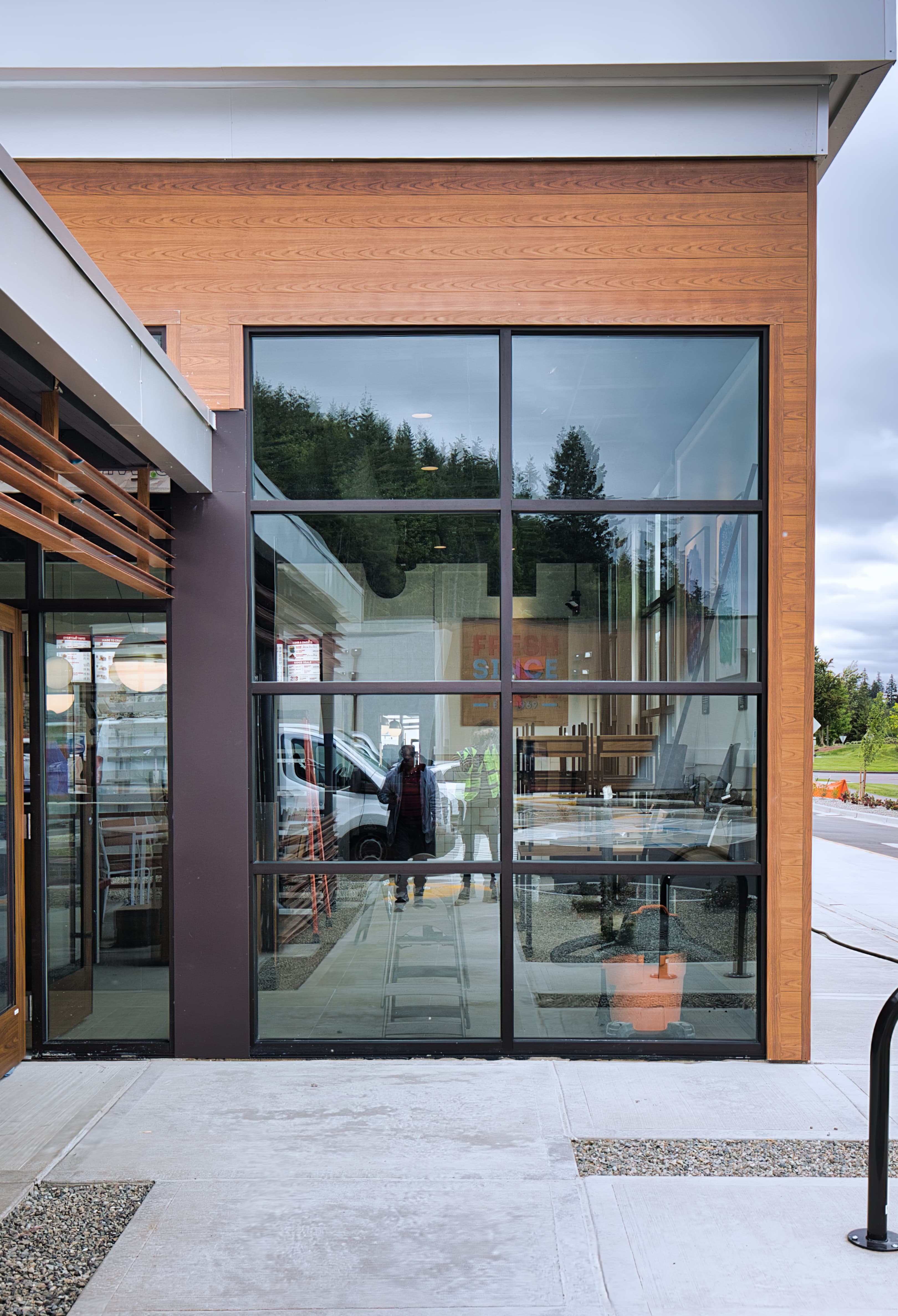 Modern building exterior with wood paneling and large windows reflecting people and evergreen trees.