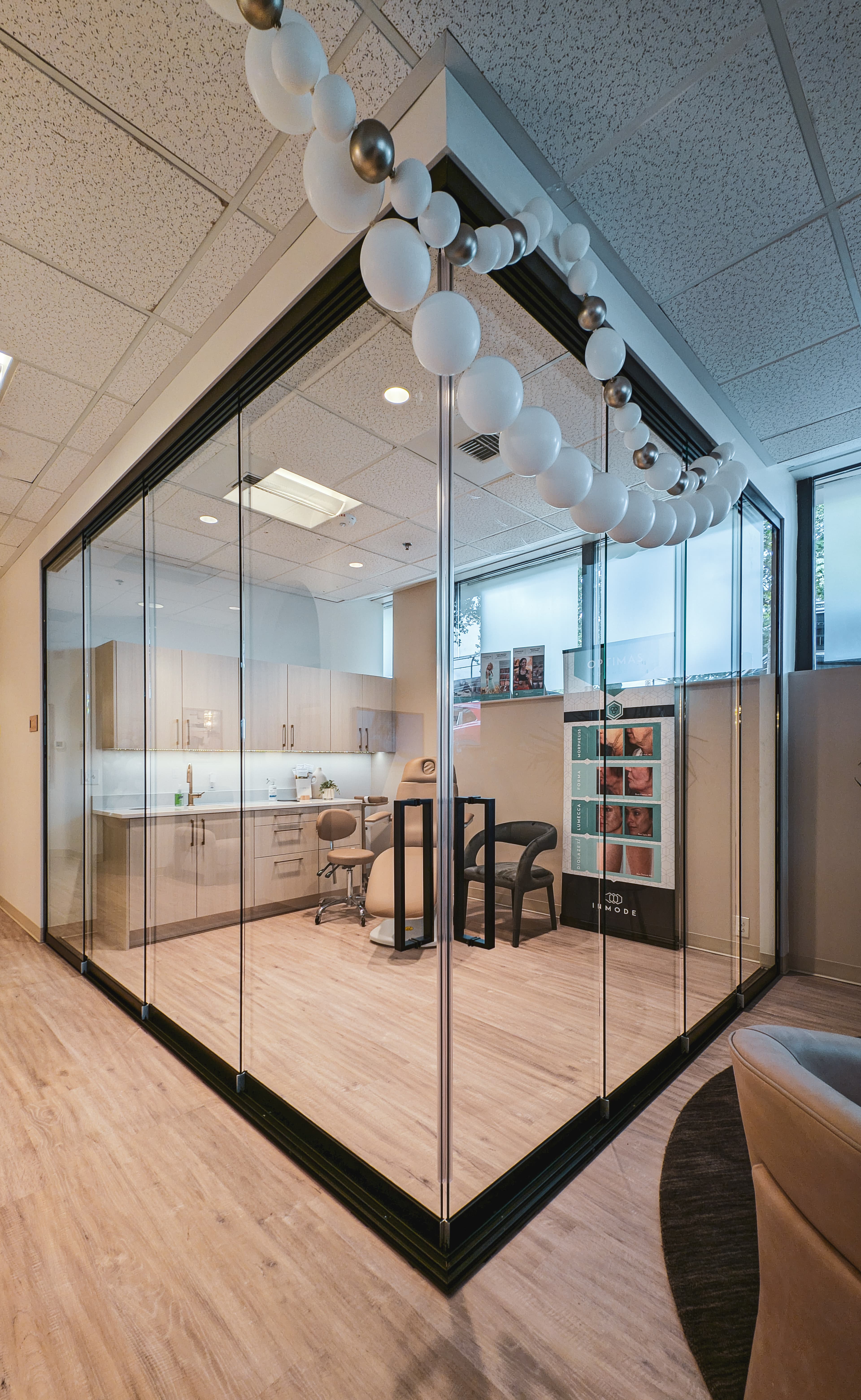 Glass-walled medical treatment room featuring a tan procedure chair, cabinetry, and festive balloon garland.